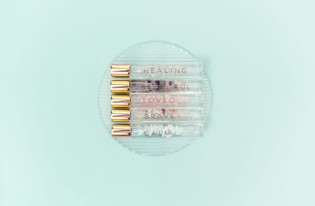 5 Crystal Roller Bottles with Essential Oil Blends and gold caps lined up on a clear glass plate with a pale aqua backdrop