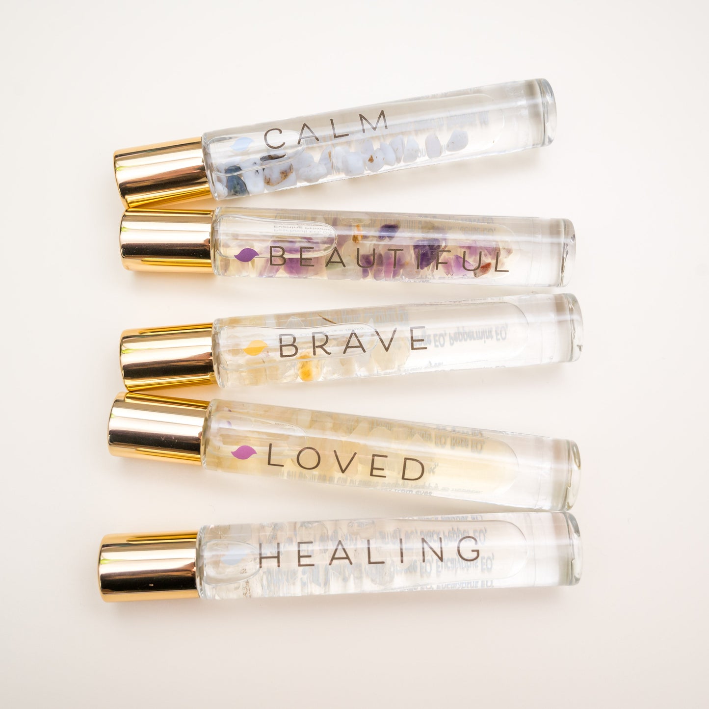 Clear Glass Roller bottles with gold caps contains essential oil blends with crystals inside called Calm, Beautiful, Brave, Loved and Healing on a white ivory background