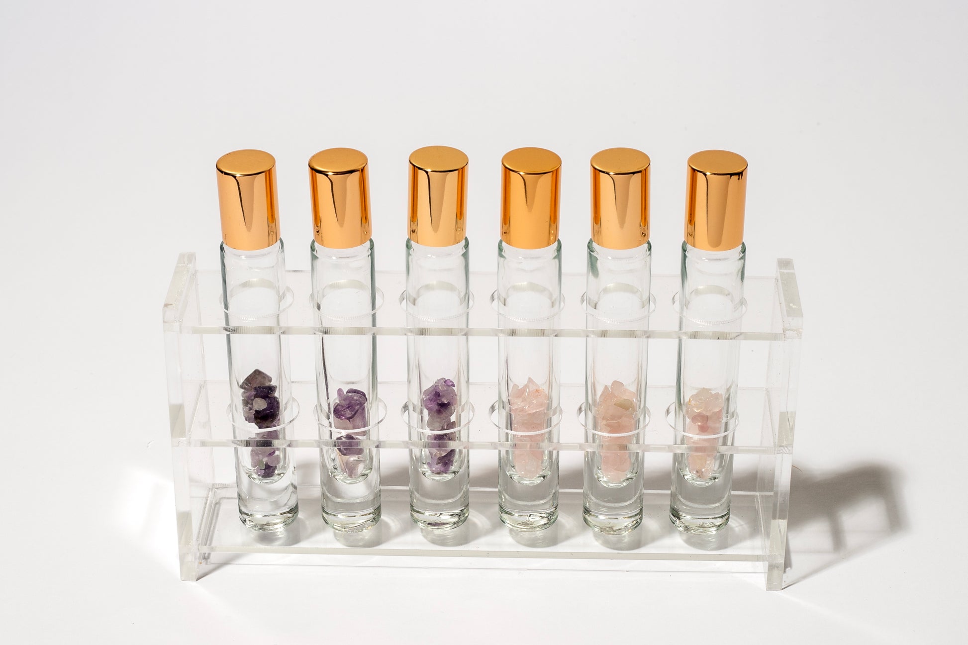 6 clear glass roller bottles in a display stand that contain amethyst and rose quartz with gold caps