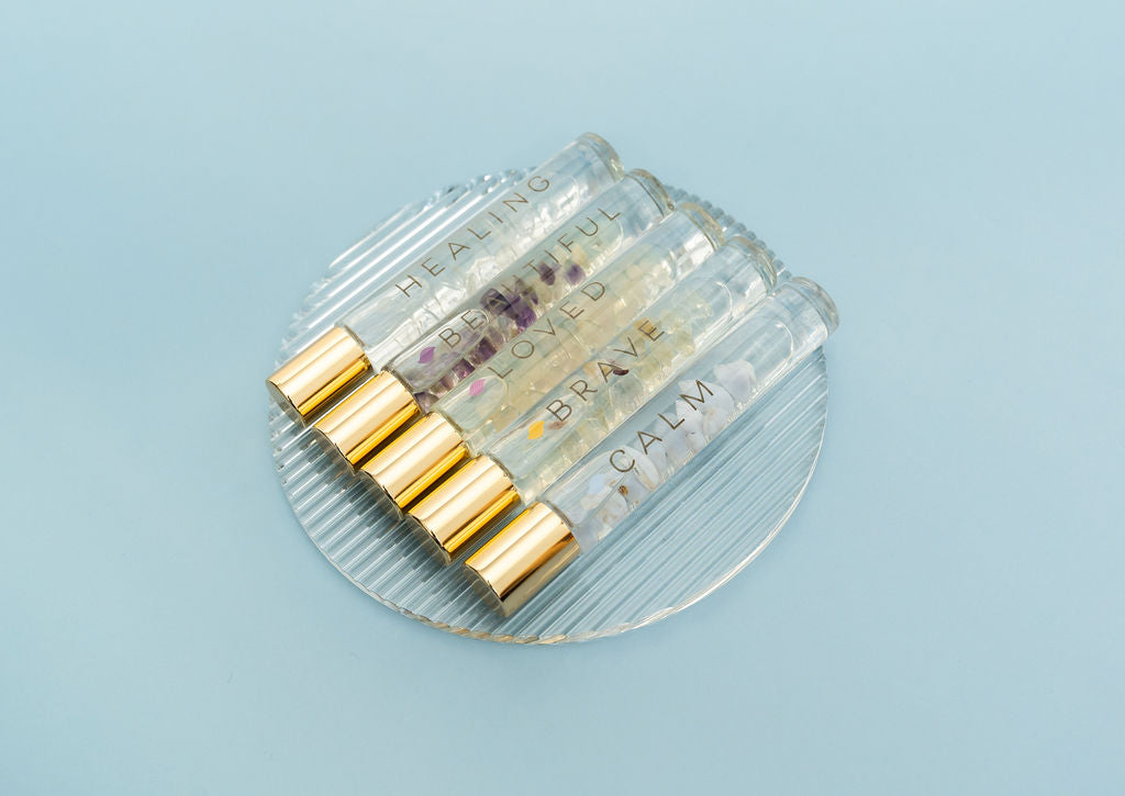 5 Crystal Essential Oil Rollers diag on a clear glass plate with gold caps on pale aqua background called Healing, Beautiful, Loved, Brave and Calm