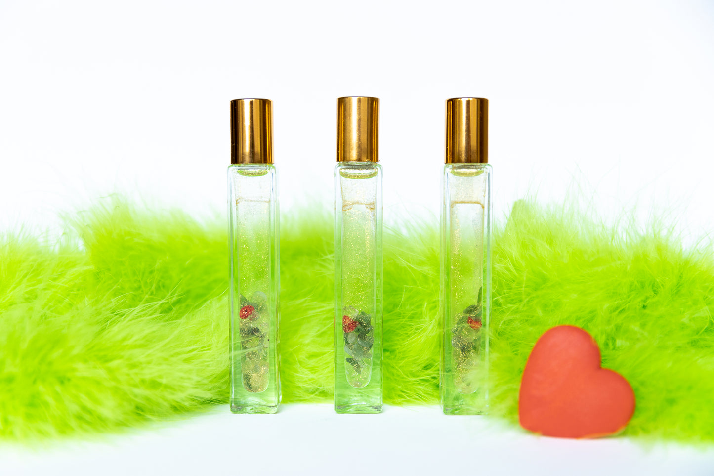 3 glass roller bottles with gold mica, green aventurine crystals and red glass heart with green fur on white background and a red grow heart