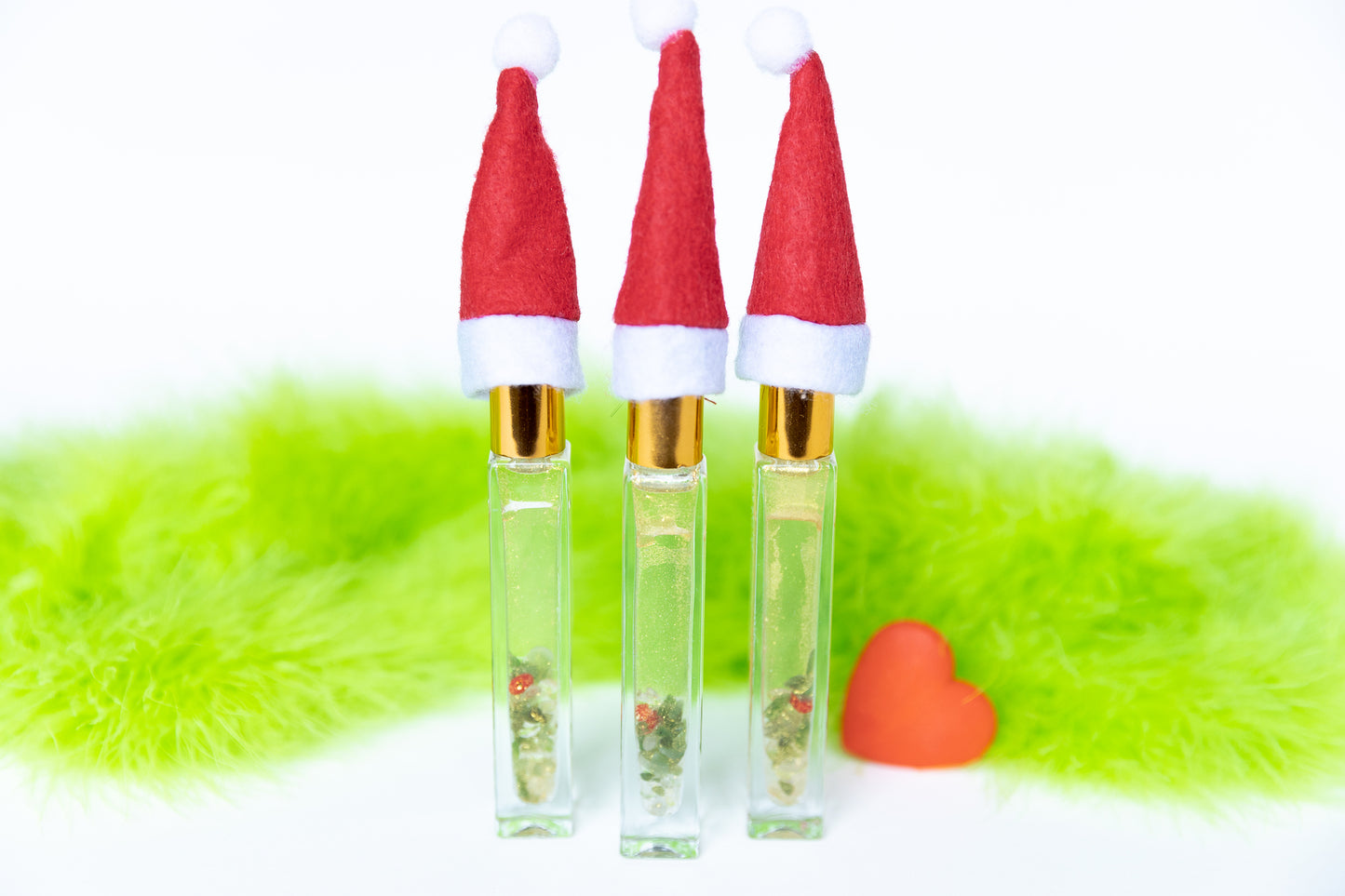 3 glass roller bottles with Santa hats that contain gold mica, green aventurine crystals and red glass heart with green fur on white background and a red grow heart