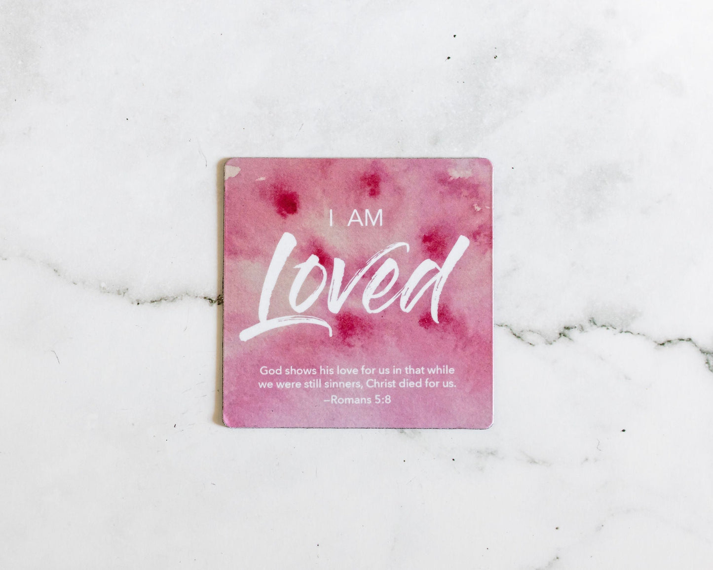 A Pink and white christian watercolor magnet that says I Am Loved with a bible verse from Romans 5:8 on a white marble background that says God shows his love for us in that while we were still sinners, Christ died for us