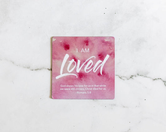 A Pink and white christian watercolor magnet that says I Am Loved with a bible verse from Romans 5:8 on a white marble background that says God shows his love for us in that while we were still sinners, Christ died for us
