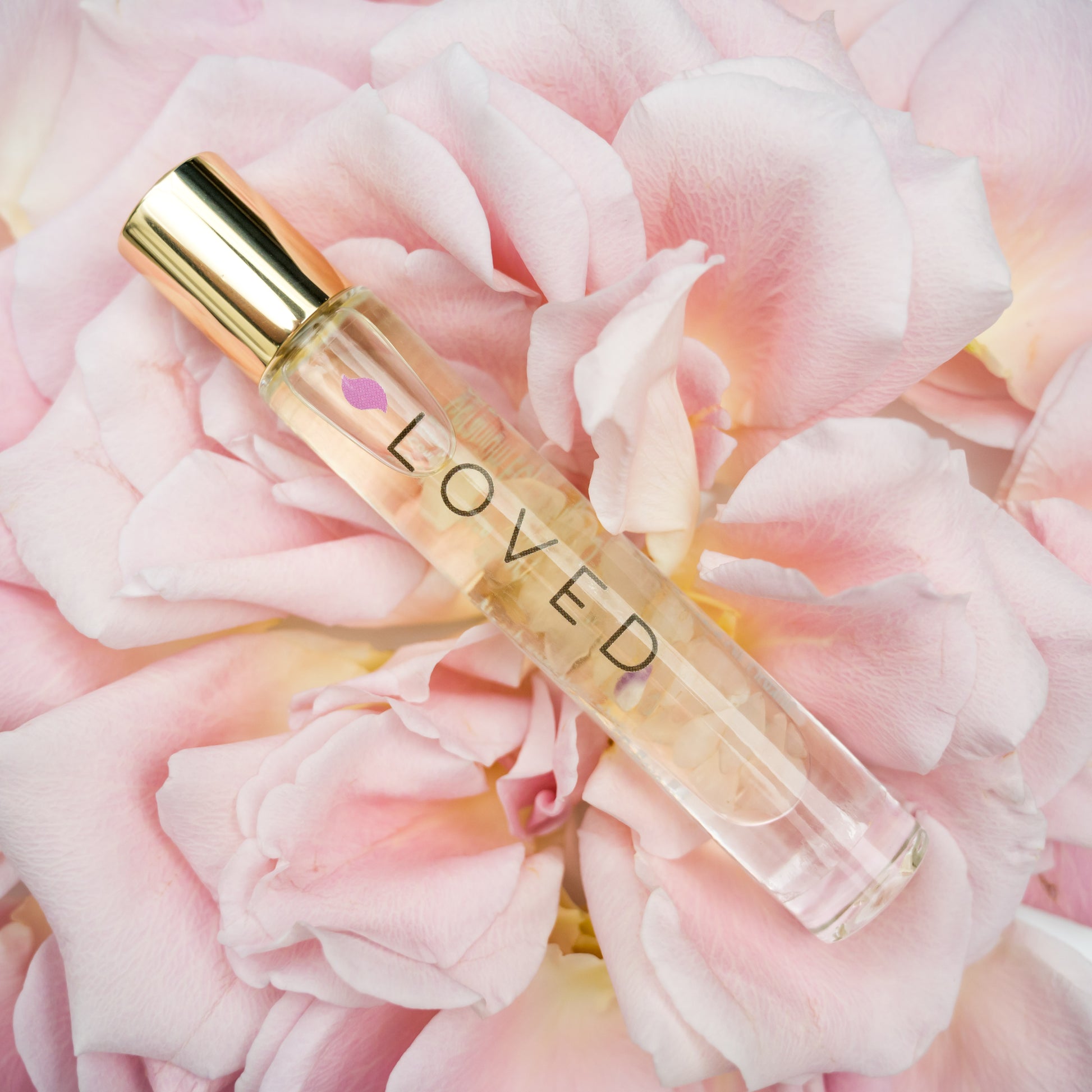 A Loved Crystal Roller Bottle Blend in glass bottle with gold cap containing rose quartz crystals laying on pink roses