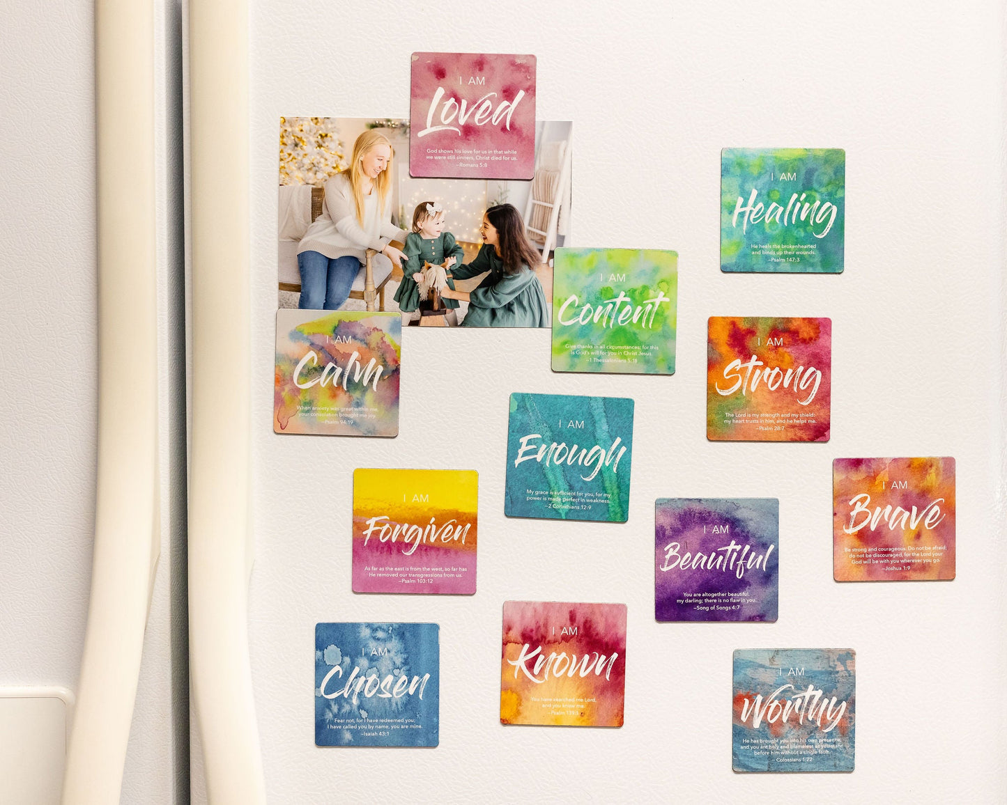 A picture of a Refrigerator door with 12 different magnets on it that each say a different affirmation word designed with water colors and holding photos to the fridge