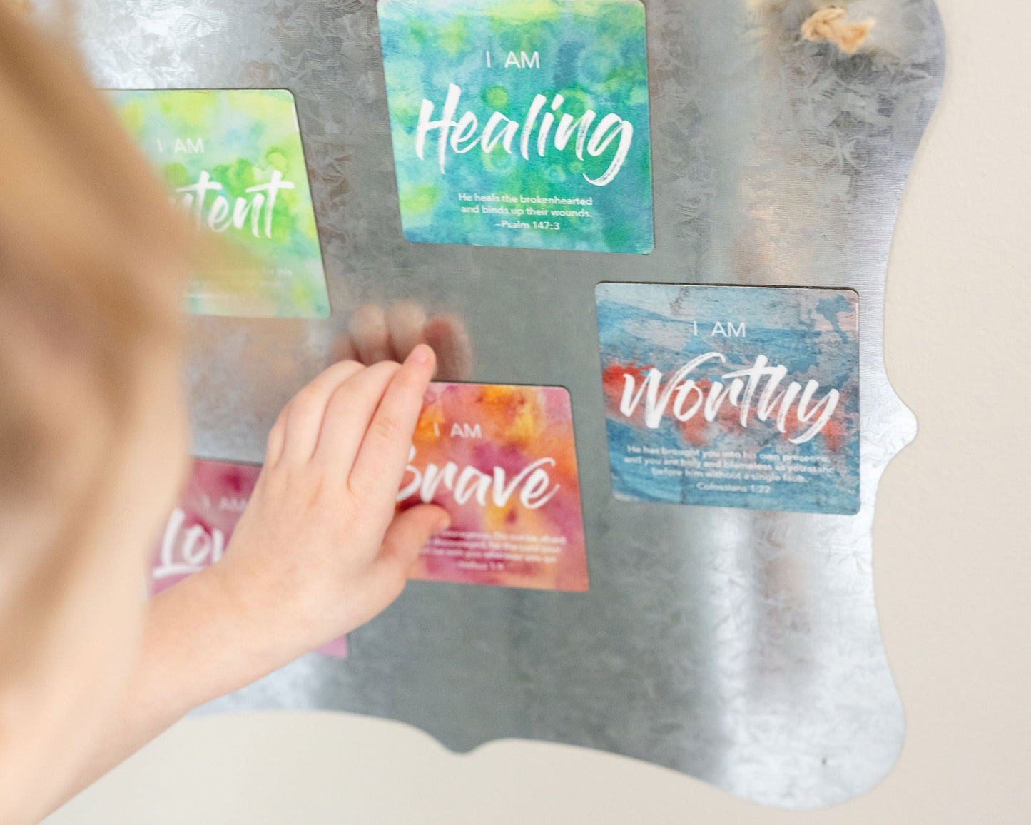 A little girls hand reaching to take a magnet that says I Am Brave that is on a hanging tin board with other colorful magnets