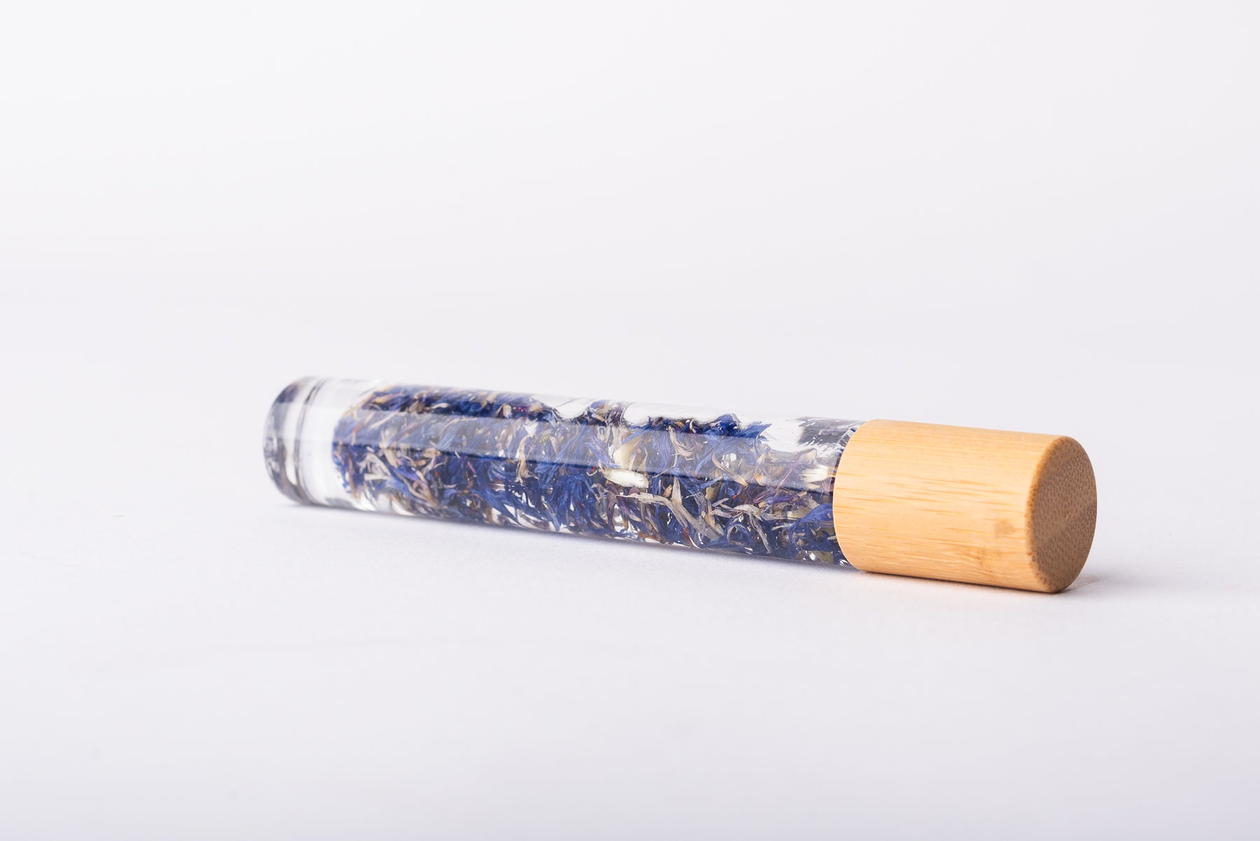 A clear glass roller bottle with Bamboo lid laying on it's side filled with organic blue cornflower buds