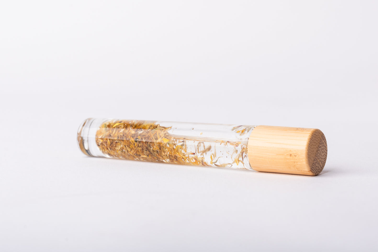 A clear glass roller bottle with Bamboo lid laying on it's side filled with organic calendula flower buds