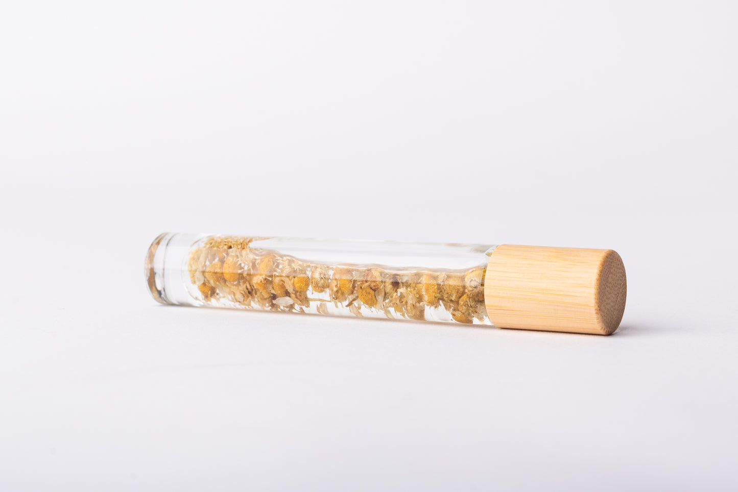 A clear glass roller bottle with Bamboo lid laying on it's side filled with organic chamomile flower buds