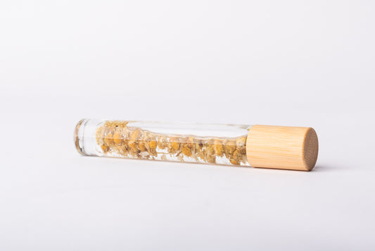 A clear glass roller bottle with Bamboo lid laying on it's side filled with organic chamomile flower buds