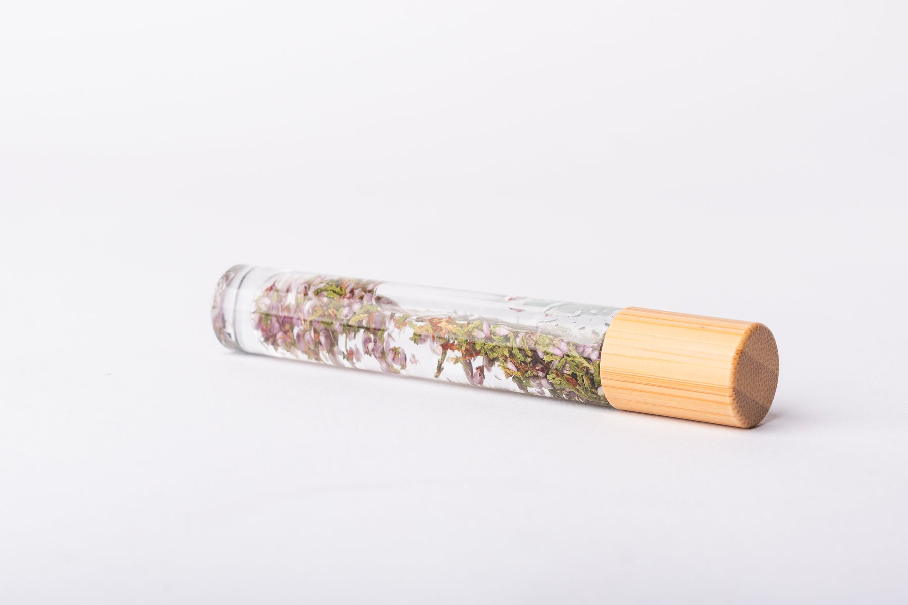 A clear glass roller bottle with Bamboo lid laying on it's side filled with organic heather flower buds