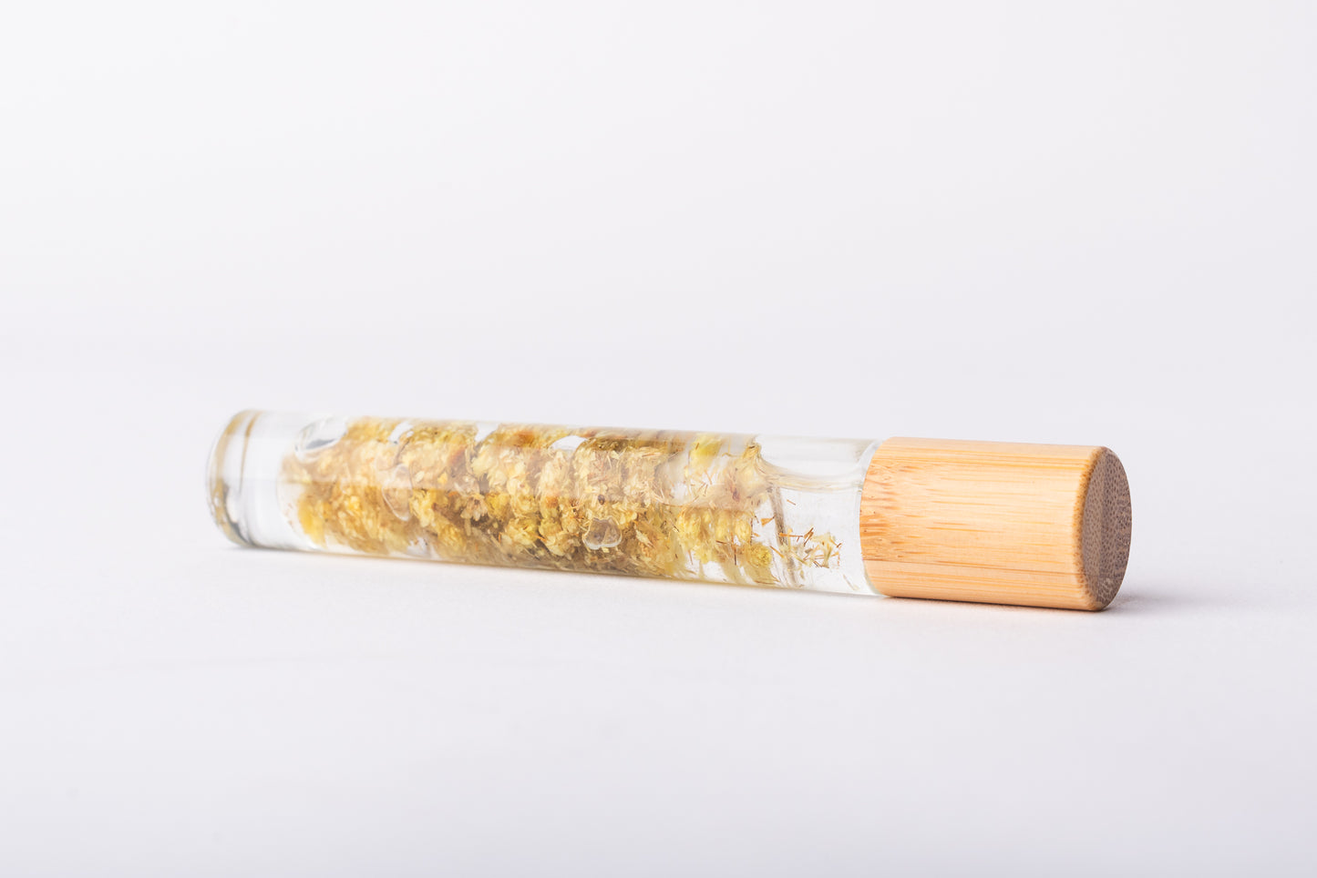 A clear glass roller bottle with Bamboo lid laying on it's side filled with organic helichrysum flower buds