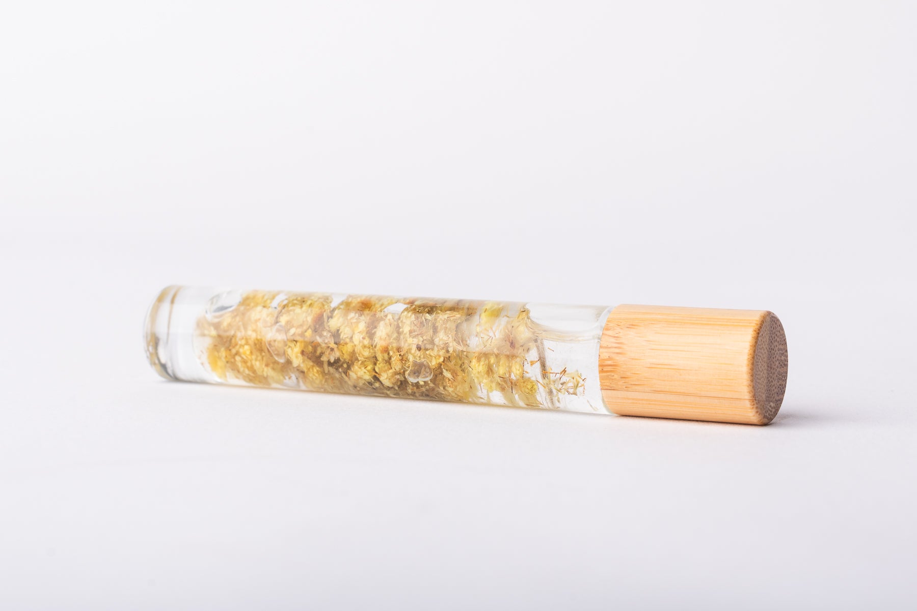 A clear glass roller bottle with Bamboo lid laying on it's side filled with organic helichrysum flower buds