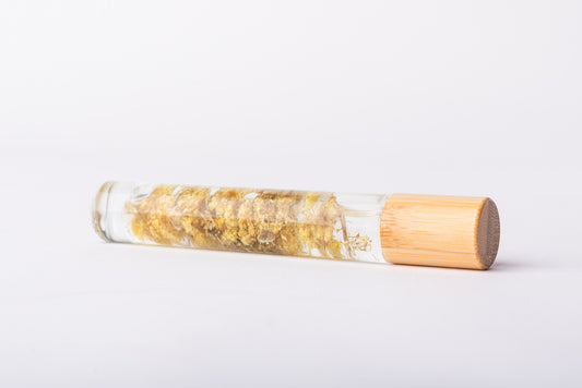 A clear glass roller bottle with Bamboo lid laying on it's side filled with organic helichrysum flower buds