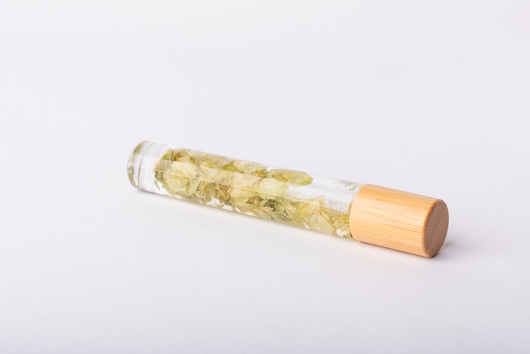 A clear glass roller bottle with Bamboo lid laying on it's side filled with organic hops leaves
