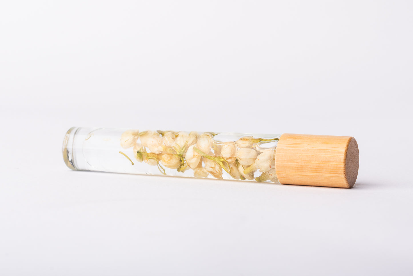 A clear glass roller bottle with Bamboo lid laying on it's side filled with organic jasmine flower buds