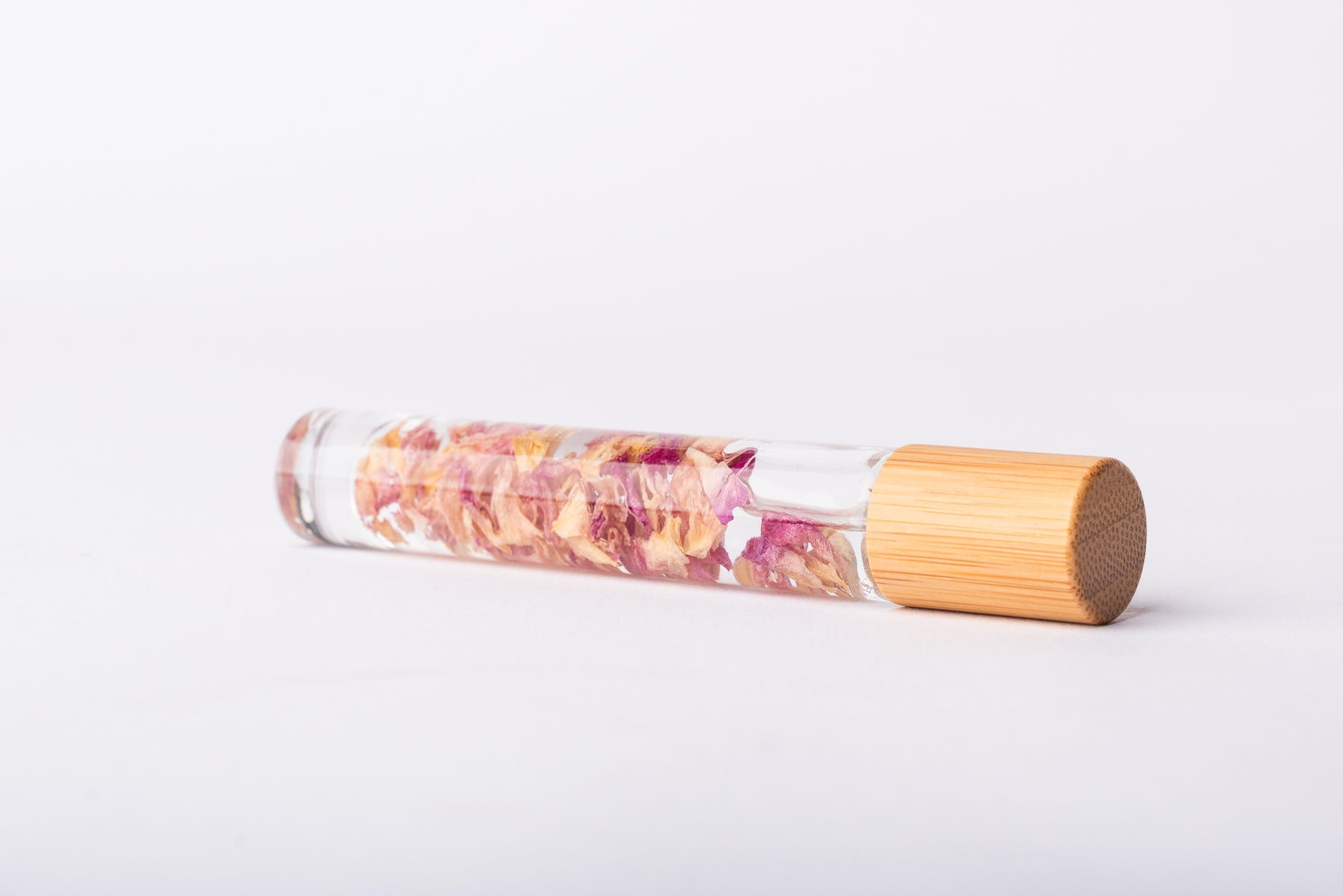 A clear glass roller bottle with Bamboo lid laying on it's side filled with organic pink rose petals