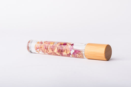 A clear glass roller bottle with Bamboo lid laying on it's side filled with organic pink rose petals