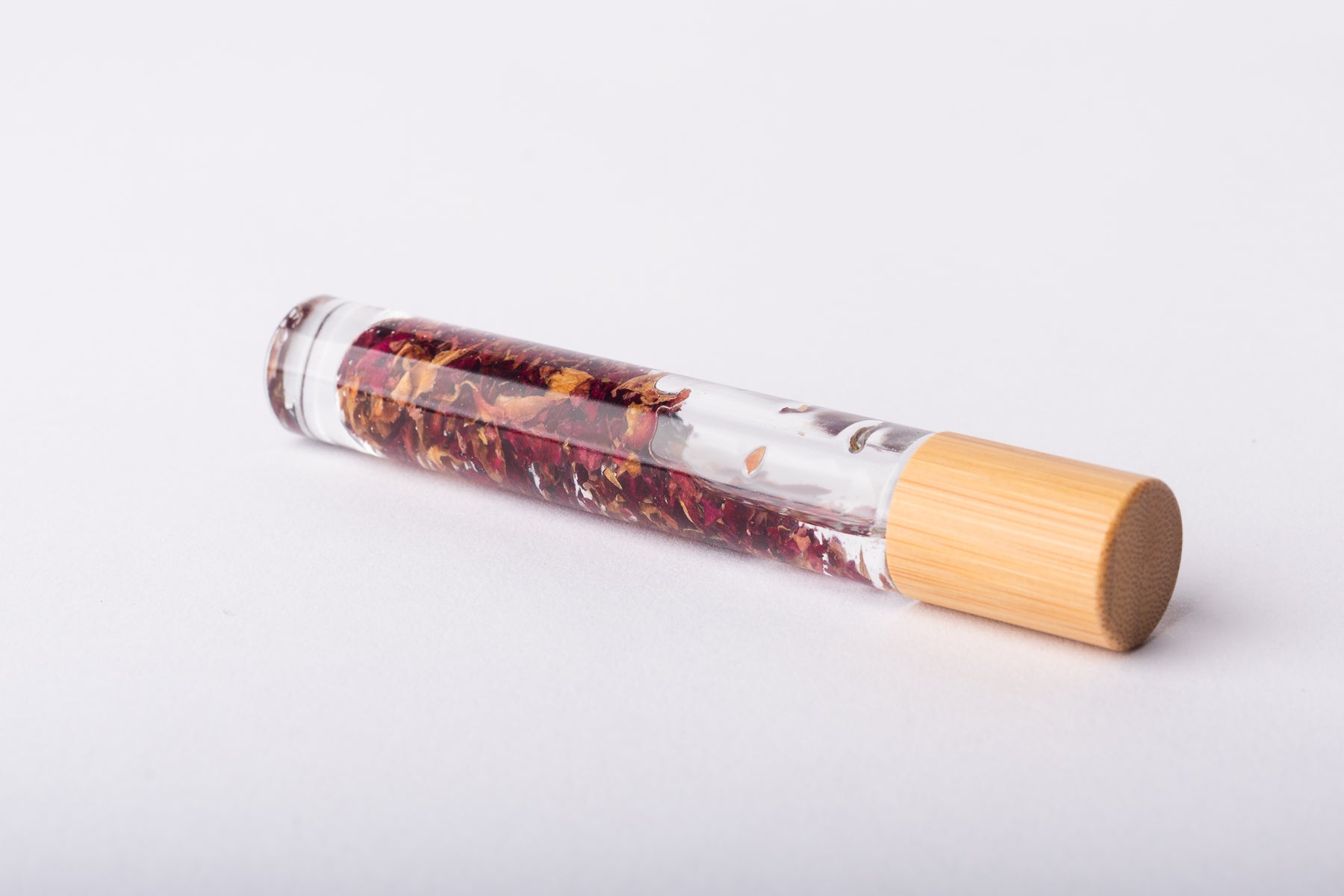 A clear glass roller bottle with Bamboo lid laying on it's side filled with organic red rose petals