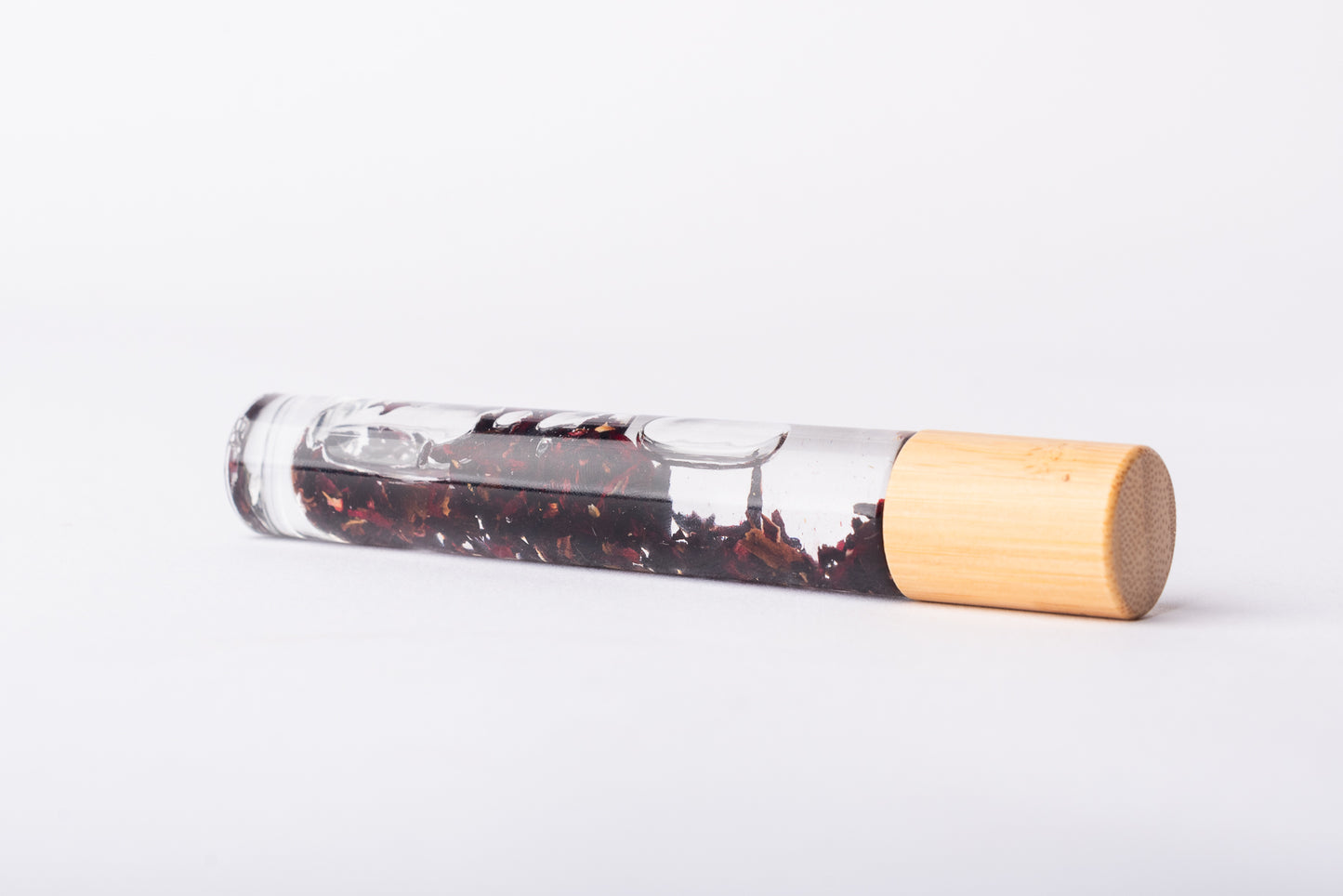 A clear glass roller bottle with Bamboo lid laying on it's side filled with organic rosehip flower buds