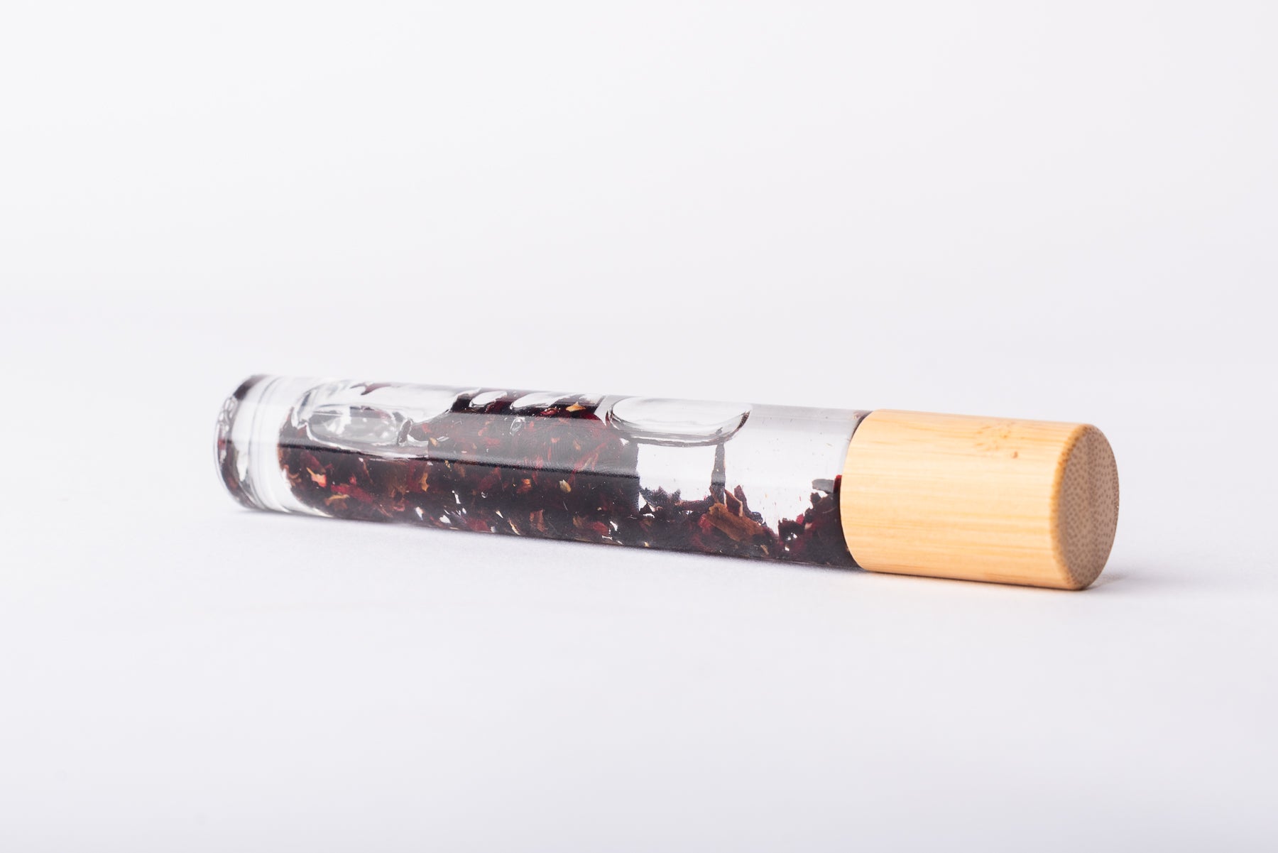 A clear glass roller bottle with Bamboo lid laying on it's side filled with organic rosehip flower buds