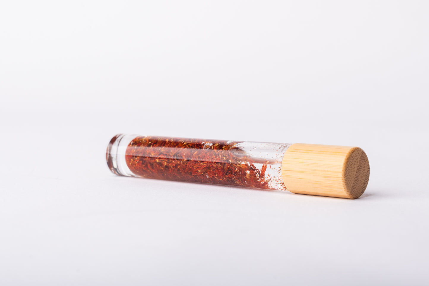 A clear glass roller bottle with Bamboo lid laying on it's side filled with organic safflower flower buds
