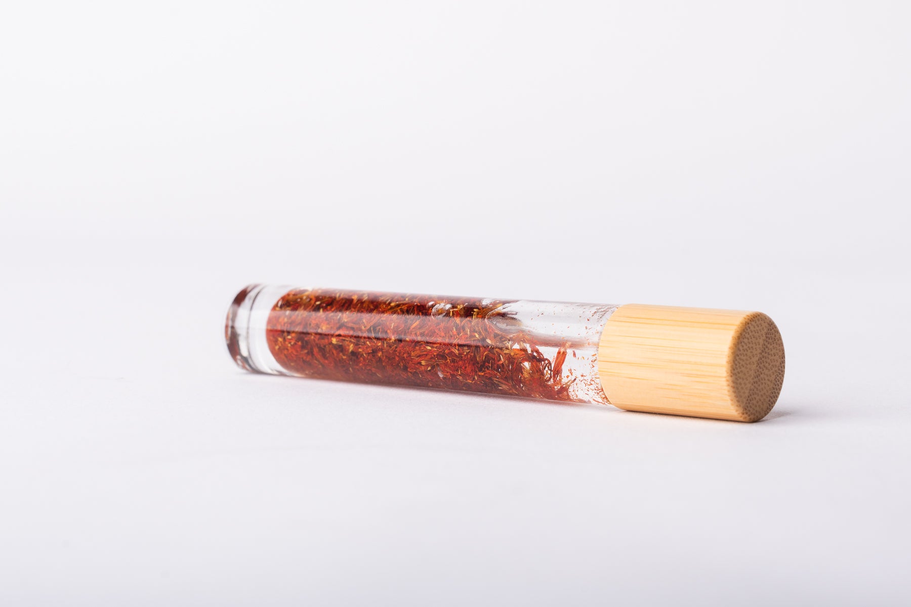 A clear glass roller bottle with Bamboo lid laying on it's side filled with organic safflower flower buds