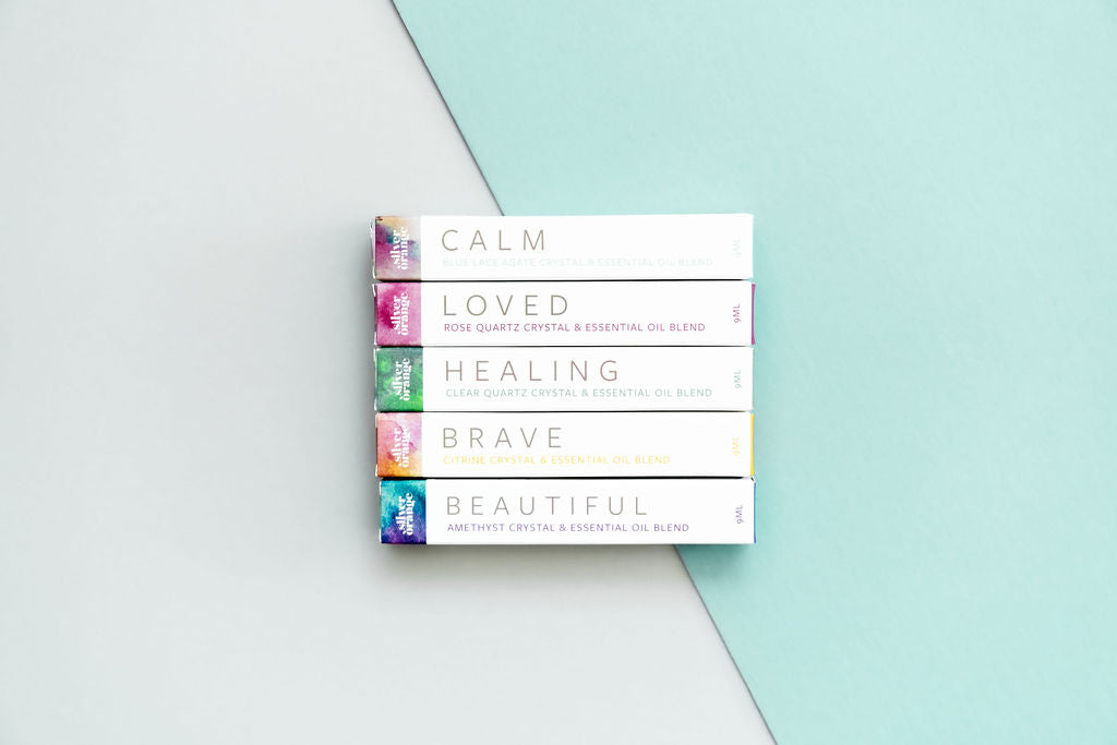5 boxes with watercolor ends that hold Crystal Essential Oil Blends in Roller Bottles on a backdrop that has half white and half aqua on a diag.