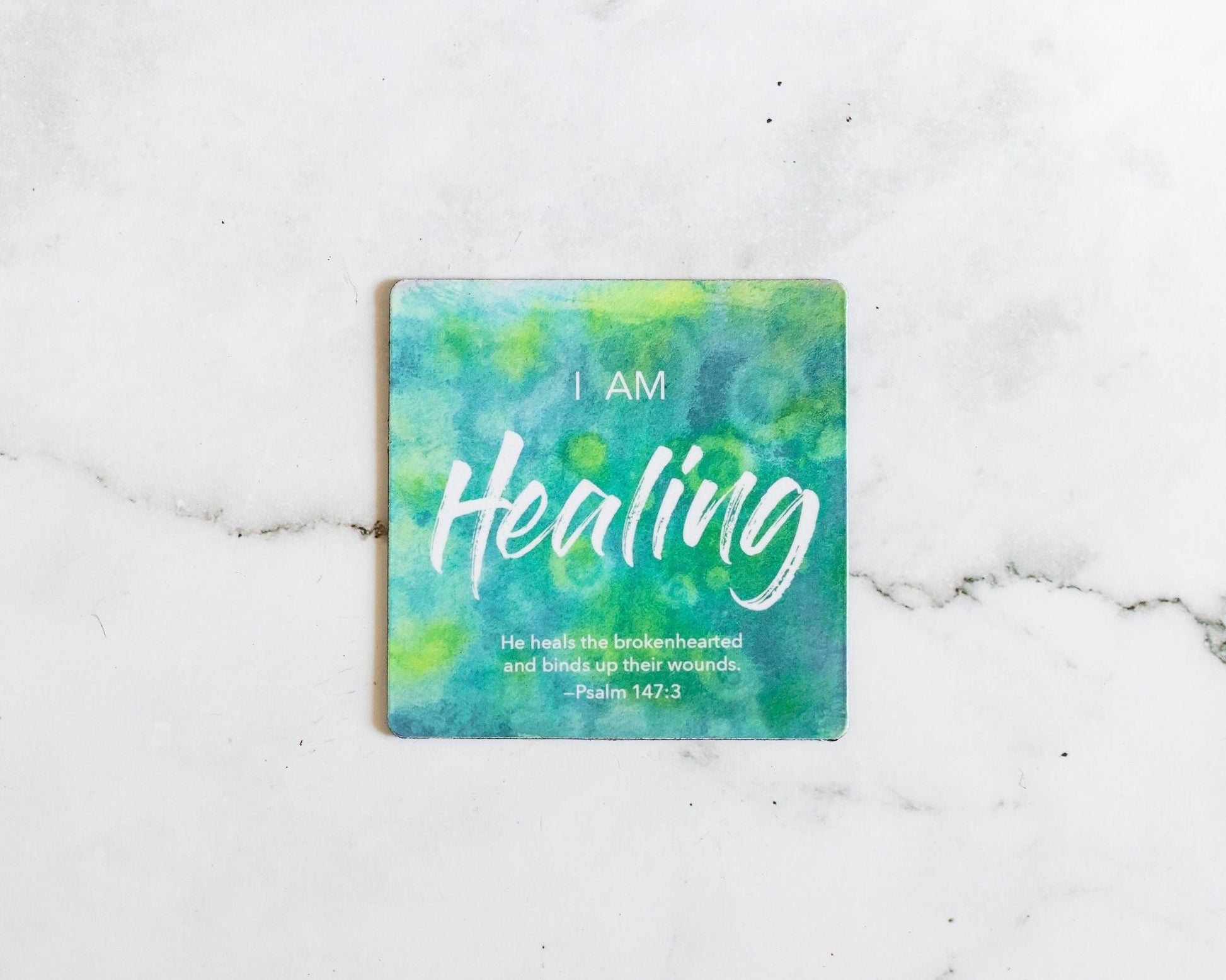 A Green and Blue magnet that says I Am Healing with a bible verse from Psalm 147:3 on a white marble background that says He heals the brokenhearted and binds up their wounds